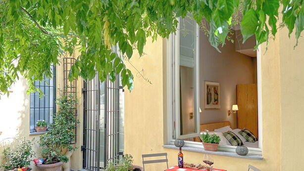 A small patio with potted plants, a table with a bottle and glass, and a window overlooking a room with a bed and a lamp. - Cirillo Luxury Suite 16 - Arco della Pace (Milano)