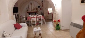 A living room with a white sofa, a small table with a vase of flowers, a dining area with a table and chairs, and an arched doorway.