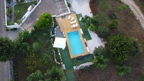 An aerial view of a property with a rectangular swimming pool, a covered patio area, and surrounding greenery.