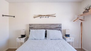 A bedroom with a bed, bedside tables, a bedside lamp, a wall-mounted sign, and a wooden shelf with decorative items. - Adventure Loft: Near Trails, Pool and Golf (Sedona)