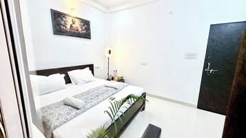 A hotel room with a bed, a nightstand, a wall-mounted light, a door, and a potted plant.