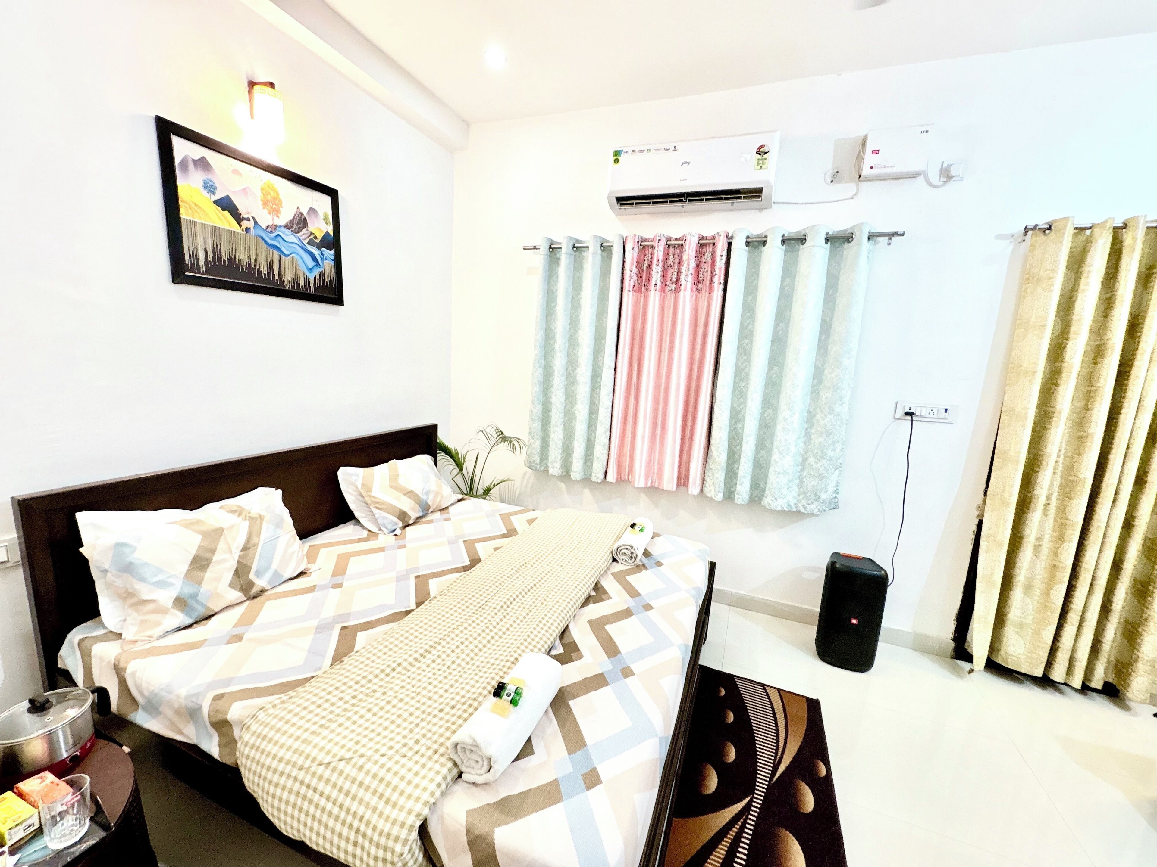 A hotel room with a bed, a wall-mounted air conditioner, a painting, and curtains.