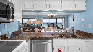 A modern kitchen with white cabinets, granite countertops, stainless steel appliances, and a view to the living area. - Emerald Towers 503 (Destin)