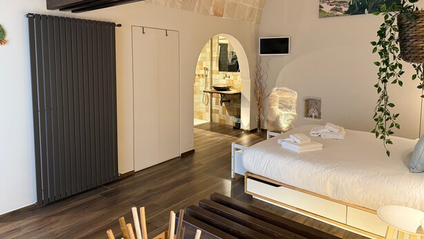 Una camera da letto con un letto, pavimento in legno, parete in pietra e vista su un bagno con lavandino e doccia.
