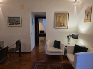 Un salon avec un canapé, un fauteuil et une petite table sur laquelle se trouve une lampe. On aperçoit un téléviseur au fond et un tableau au mur.