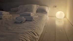 Una cama bien hecha con sábanas y almohadas blancas, una lámpara de pared redonda y un enchufe en la pared.
