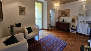 Un salón espacioso con techo alto, suelo de madera y un tapiz grande. Hay dos sillones blancos, un aparador de madera y un cuadro en la pared.