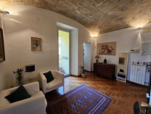 Un salon spacieux avec un plafond haut, un plancher en bois et un grand tapis. On y trouve deux fauteuils blancs, un meuble en bois et un tableau accroché au mur.