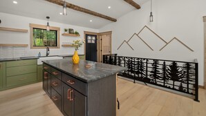 A modern kitchen with a central island, green cabinets, and a black countertop. There is a window with a view of trees, a black door, and a decorative wall panel. - Riverside Lodge, a Modern Adirondack Getaway (Boonville)