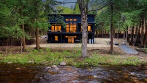 A modern two-story cabin surrounded by a forest, with a stream in the foreground. - Riverside Lodge, a Modern Adirondack Getaway (Boonville)