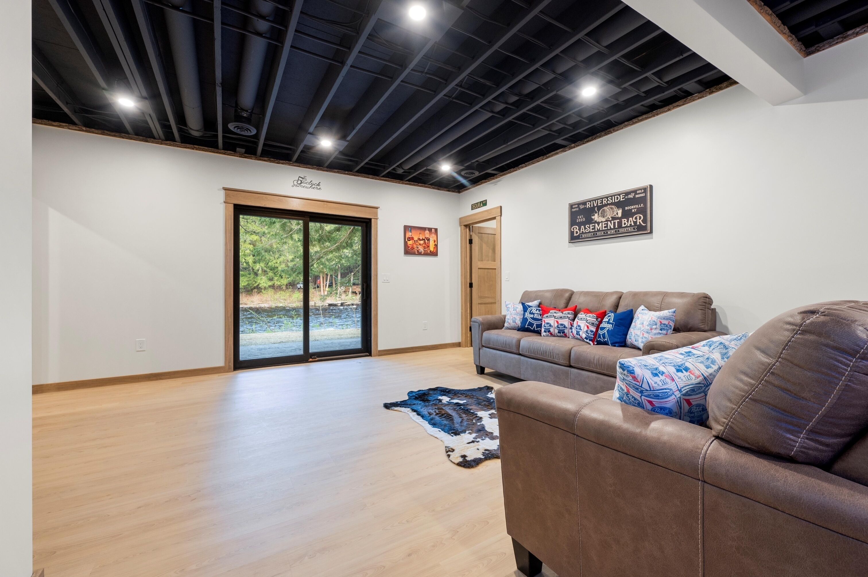 A spacious living area with a large sliding glass door leading to a balcony, a brown leather sofa with patterned cushions, and a wooden floor.