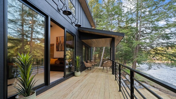 A modern house with a wooden deck, large windows, and a view of a forest and lake. - Riverside Lodge, a Modern Adirondack Getaway (Boonville)