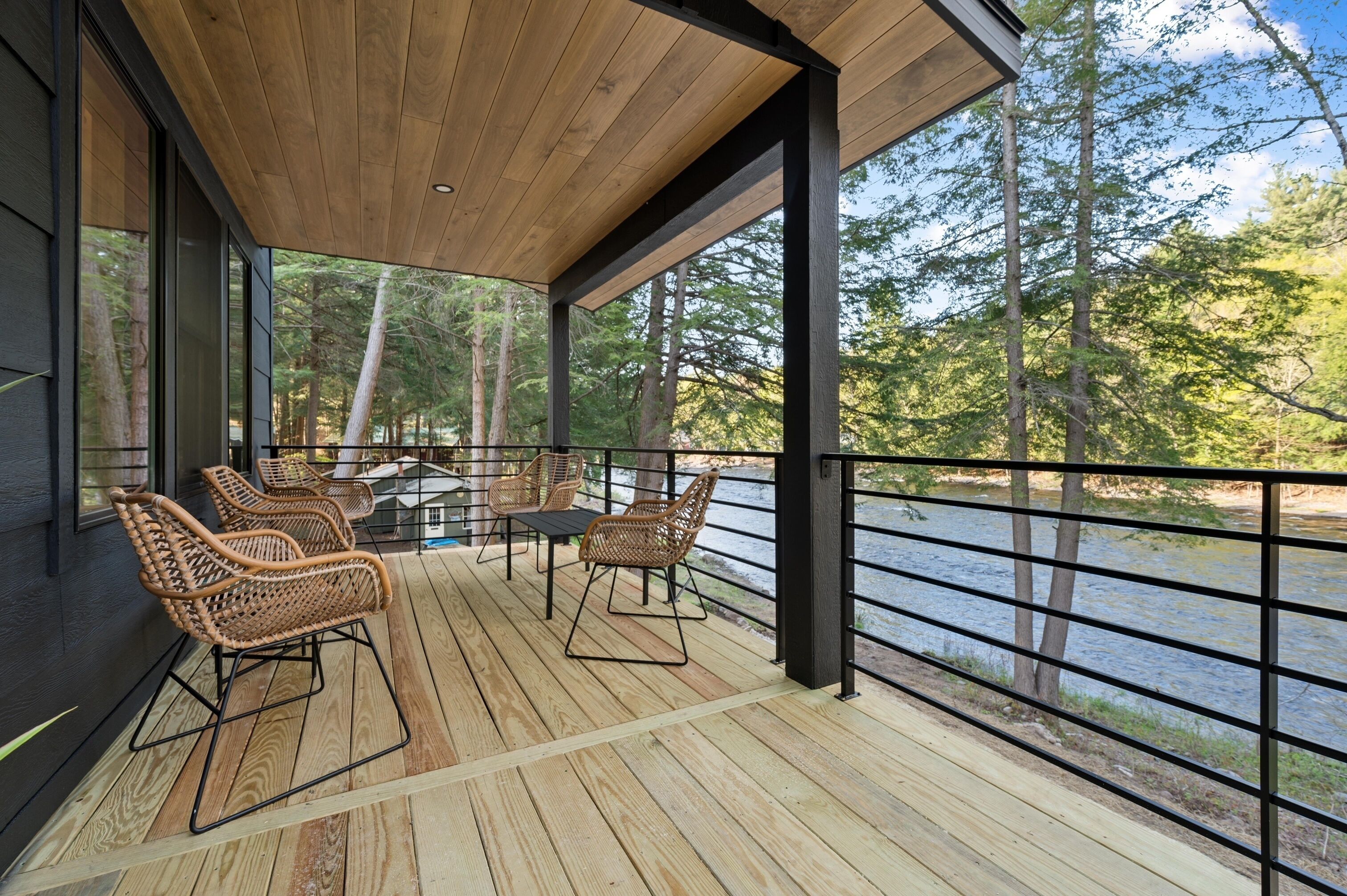 Een overdekt terras buiten met houten vloer, rieten stoelen en een tafel.