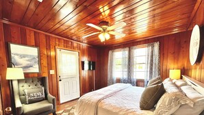 Une chambre à coucher aux murs lambrissés, avec un lit, une chaise dotée d’un coussin, un ventilateur de plafond et une porte équipée de stores.