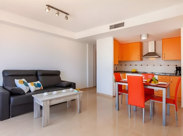 A modern living room with a sofa and coffee table, a dining area with a table and chairs, and a kitchen with orange cabinets.