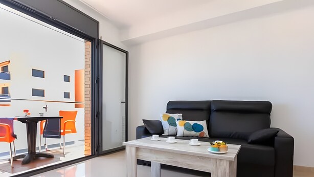 A modern living room with a black sofa, a coffee table, and a glass door leading to an outdoor area with orange chairs. - Apartment 'Tropikana I' with Shared Pool, Wi-Fi and Air Conditioning (Miami Platja)