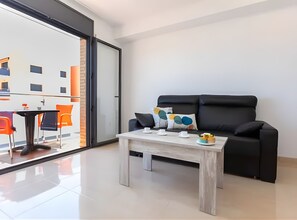 Un salon moderne avec un canapé noir, une table basse et une porte vitrée donnant sur un espace extérieur agrémenté de chaises orange.