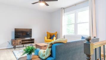 Un salon comprenant une télévision, un fauteuil jaune, un canapé bleu, une table basse en bois et un ventilateur de plafond.