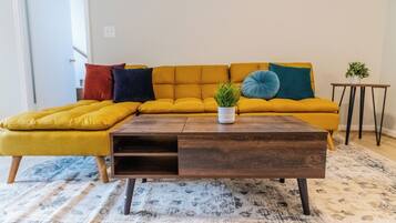 Un salon avec un canapé d’angle jaune moutarde, une table basse en bois et un tapis à motifs.