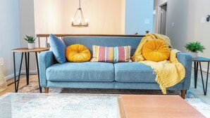 A living room with a blue sofa, yellow and multicolored pillows, a wooden coffee table, and a hanging light fixture. - Charming 4-bedroom house with AC and WiFi in Winston-Salem (Winston-Salem)