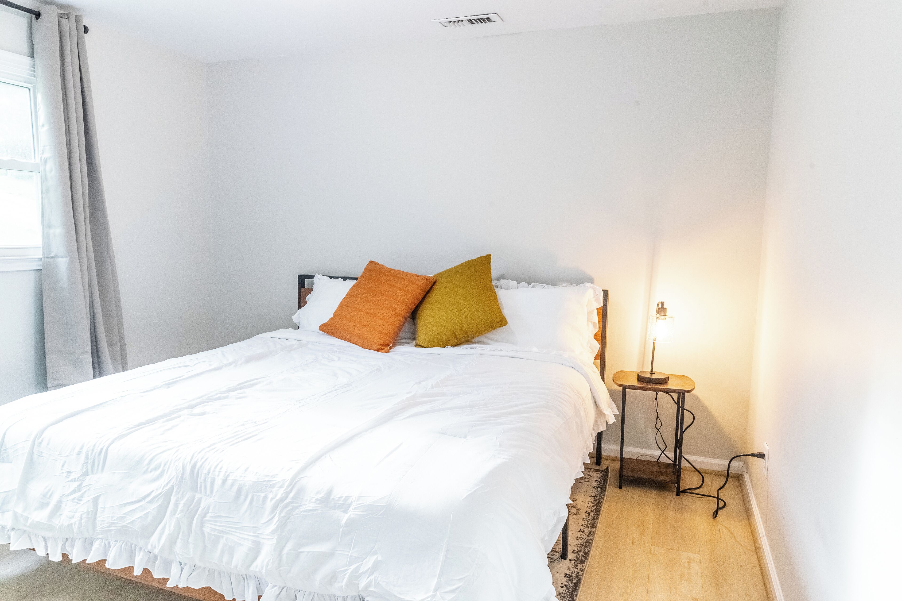 A neatly made bed with white bedding and two pillows, a bedside table with a lamp, and a window with curtains.