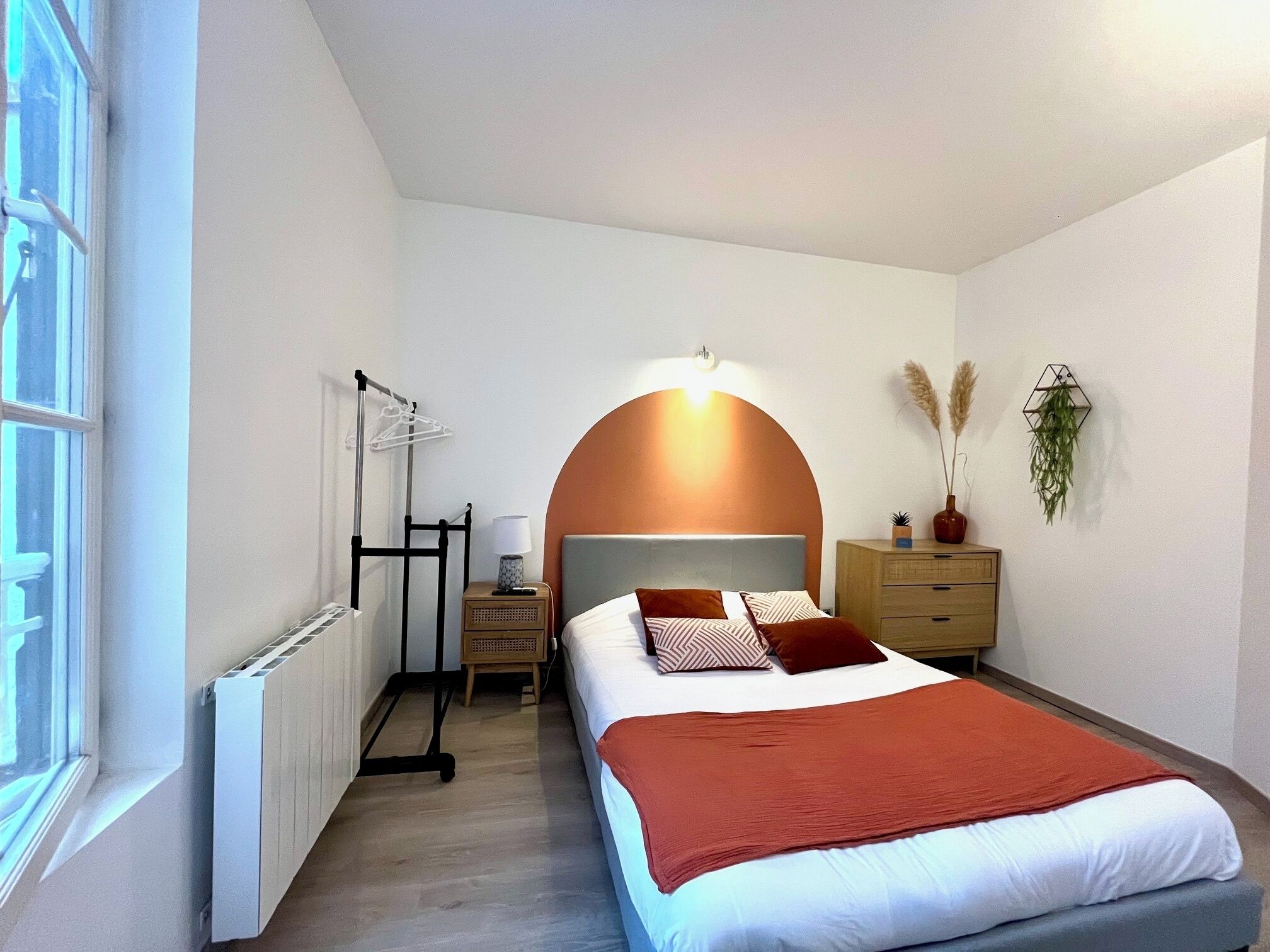 Een slaapkamer met een bed, een radiator, een nachtkastje, een lamp en een plant aan de muur.