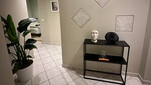 Un pasillo con una planta en maceta, un estante de metal negro con un busto y un objeto decorativo, y un suelo de baldosas.