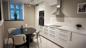 Una cocina moderna con armarios blancos, horno empotrado y campana extractora de acero inoxidable. Hay una mesa de comedor con sillas y una planta en el alféizar de la ventana.