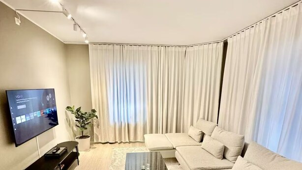 Una sala de estar moderna con un sofá seccional, un televisor de pantalla plana, una mesa de centro y un ventanal con cortinas.