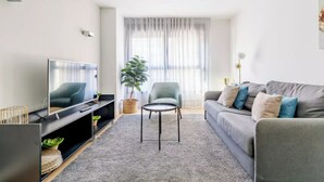Ein modernes Wohnzimmer mit einem Flachbildfernseher, einem grauen Sofa mit Kissen, einem kleinen runden Tisch und einer Zimmerpflanze.
