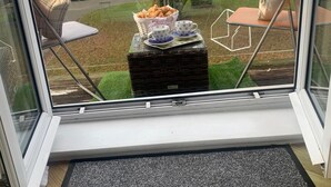 Une vue par la fenêtre sur une pièce où l’on aperçoit un panier en osier rempli de pâtisseries et un service à thé posé sur une petite table.