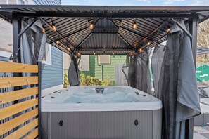 Un jacuzzi installé sous une terrasse couverte, avec une clôture en bois et une serre en arrière-plan.