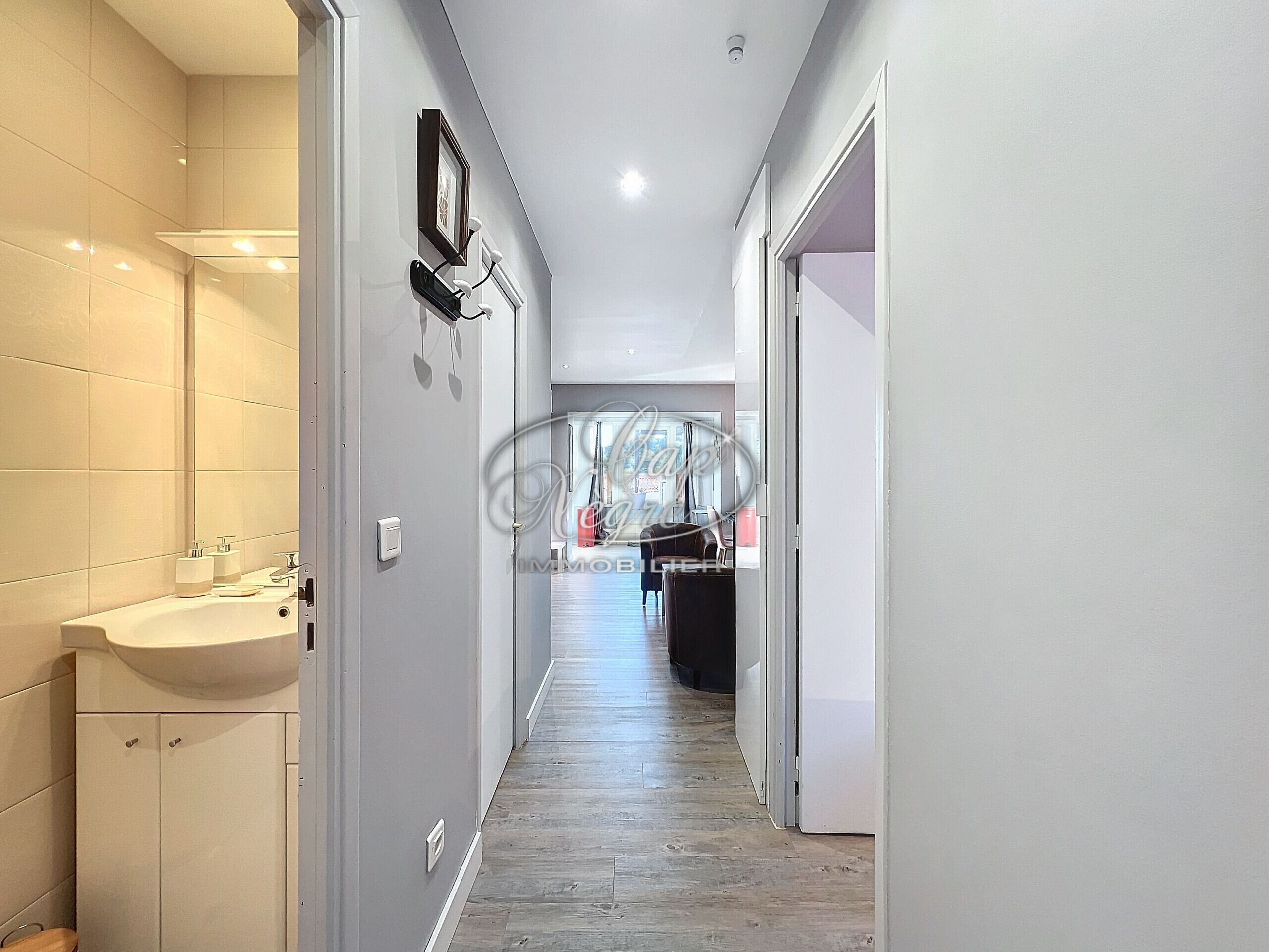 Un couloir étroit, avec une salle de bain sur la gauche, comprenant un lavabo et des placards. Le couloir est doté d’un plancher en bois et d’un luminaire au plafond.