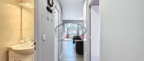 Un couloir étroit, avec une salle de bain sur la gauche, comprenant un lavabo et des placards. Le couloir est doté d’un plancher en bois et d’un luminaire au plafond.