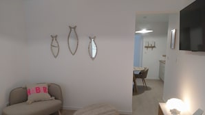 Un salon avec un canapé, une table ronde en bois et un fauteuil poire. Il y a trois miroirs fixés au mur et un téléviseur accroché au mur.