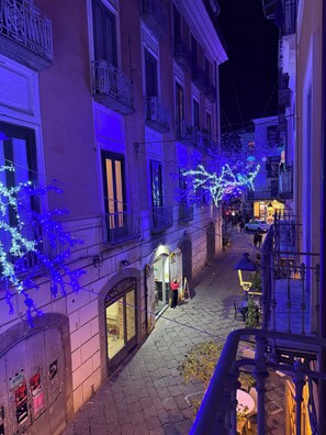 Une rue étroite, bordée d’immeubles illuminés de lumières bleues, avec une devanture de magasin bien visible et un petit espace extérieur aménagé avec des sièges.