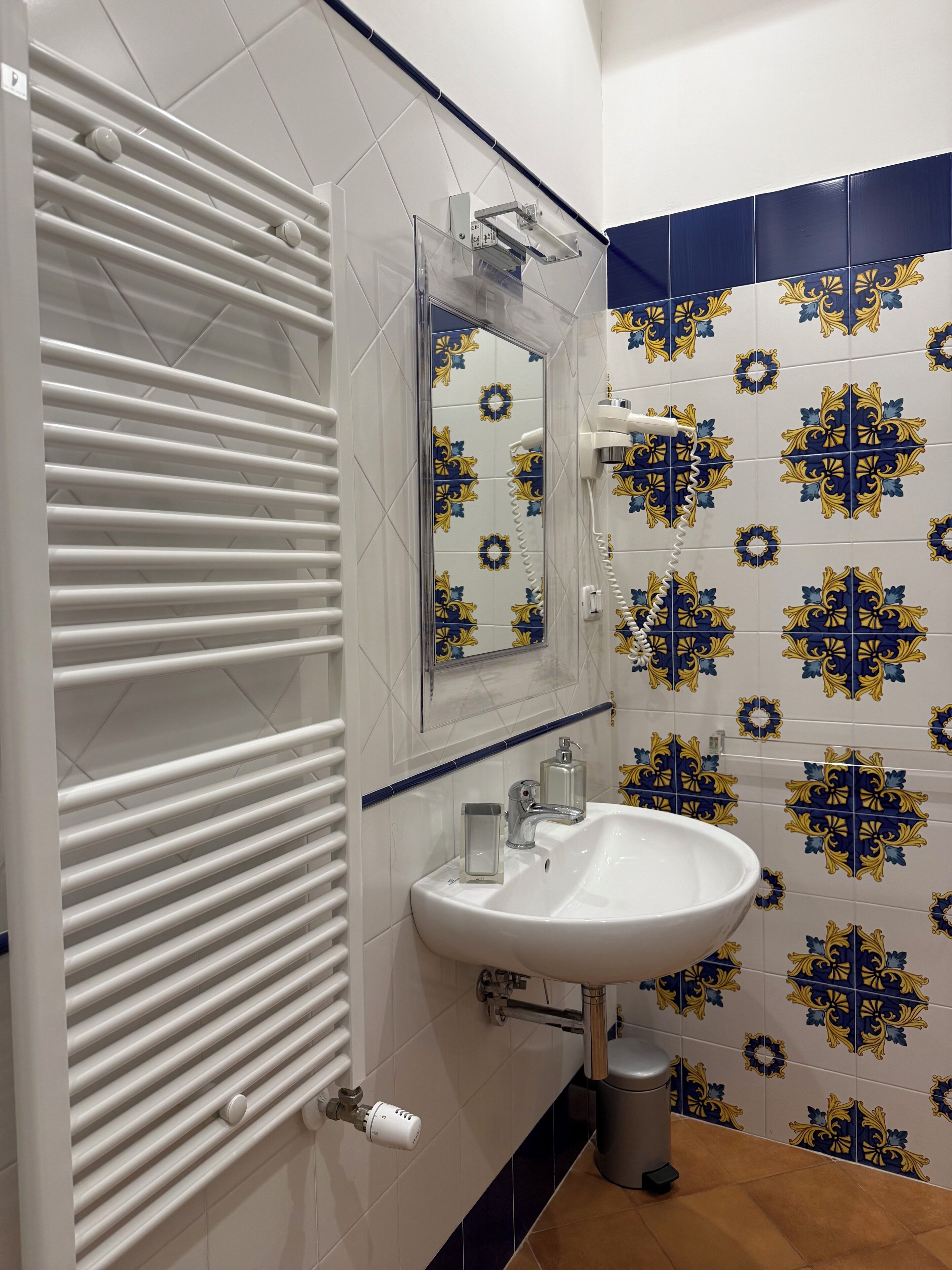 A bathroom with a white radiator, a sink with a mirror above it, and decorative blue and yellow tiles on the walls.