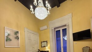 A bedroom with a bed, a chandelier, a door, a chair, a TV, and a window with a view. - Palazzo Dogana Room & Suite (Salerno)