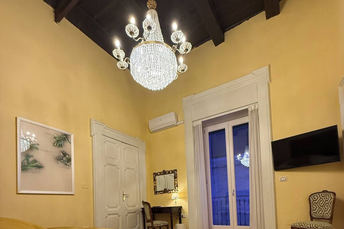 A bedroom with a bed, a chandelier, a door, a chair, a TV, and a window with a view.