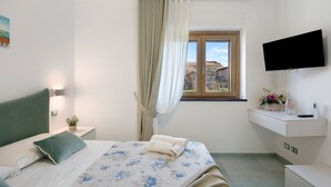 Een slaapkamer met een bed, een aan de muur bevestigde televisie, een raam met gordijnen en een klein nachtkastje met een plant.