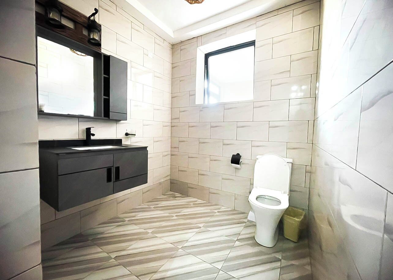 A modern bathroom with a tiled floor, a white toilet, a black vanity with a mirror, and a window providing natural light.