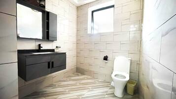 A modern bathroom with a tiled floor, a white toilet, a black vanity with a mirror, and a window providing natural light.