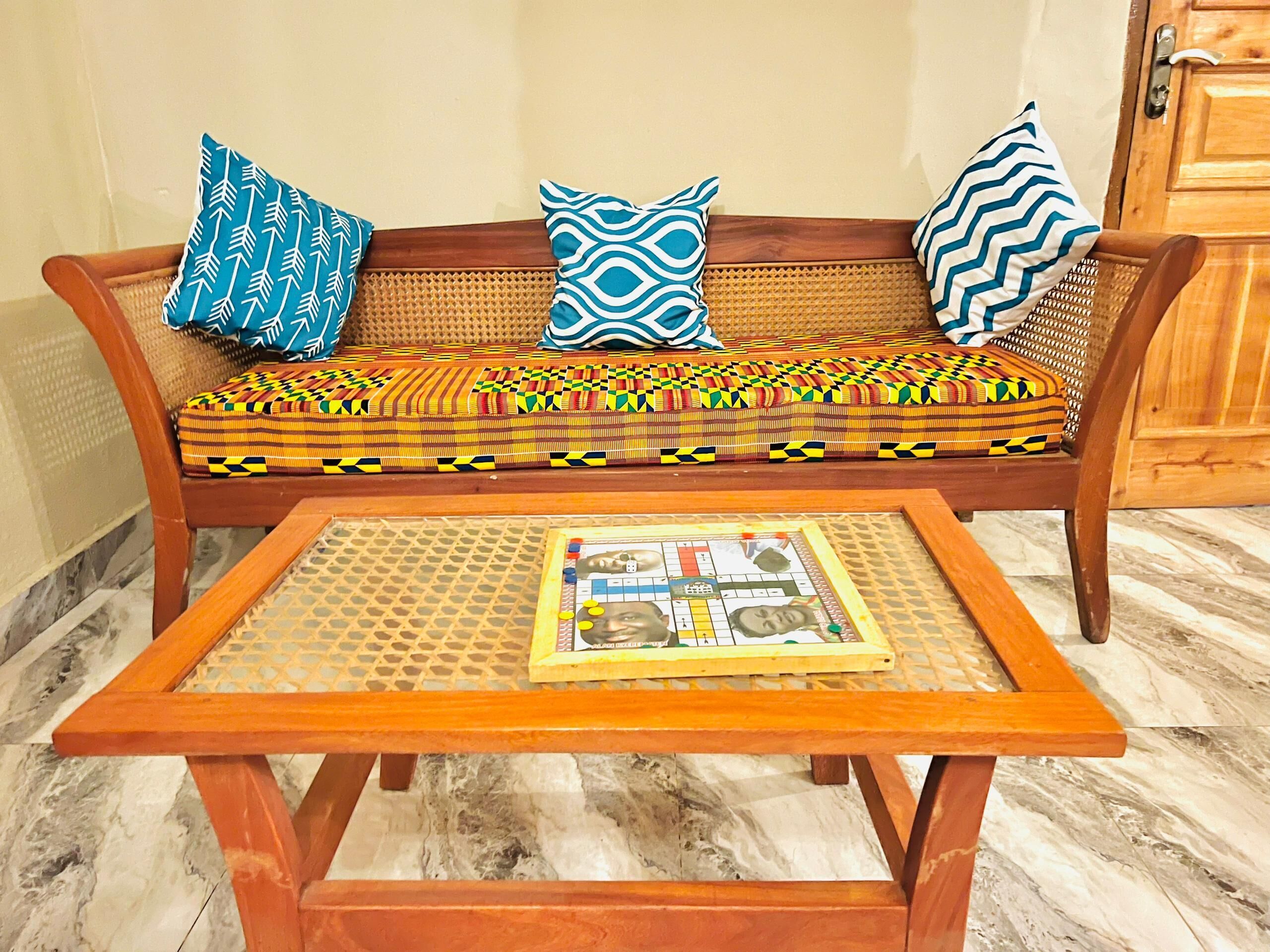 Een houten bank met gekleurde kussens en een geweven zitting, een houten salontafel met een tijdschrift, en een houten deur op de achtergrond.