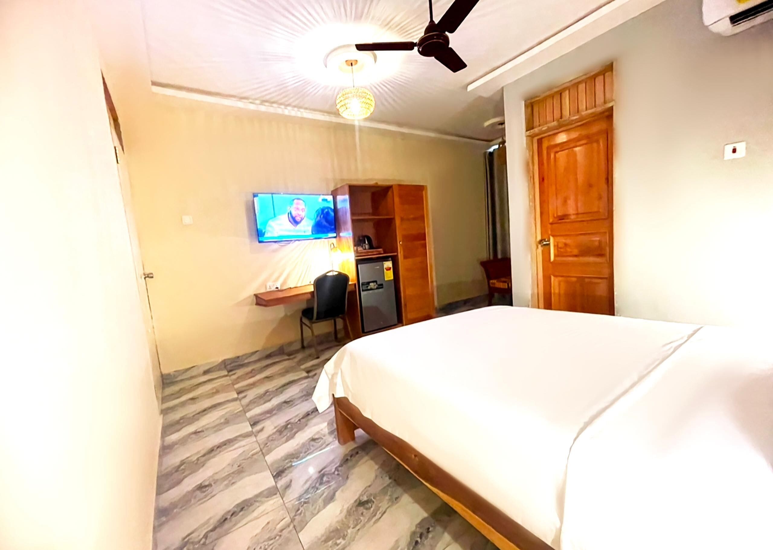 Una habitación de hotel con una cama, un escritorio con silla, un televisor, un ventilador de techo y una puerta de madera.
