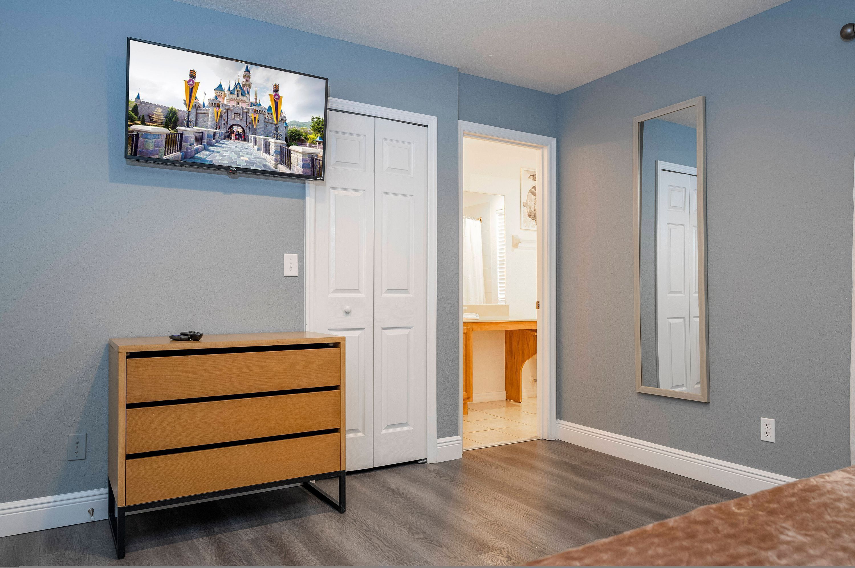 A bedroom with a wooden dresser, a television mounted on the wall, and a door leading to another room.
