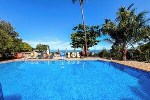 A clear blue pool with lounge chairs and umbrellas, surrounded by palm trees and a view of the ocean.