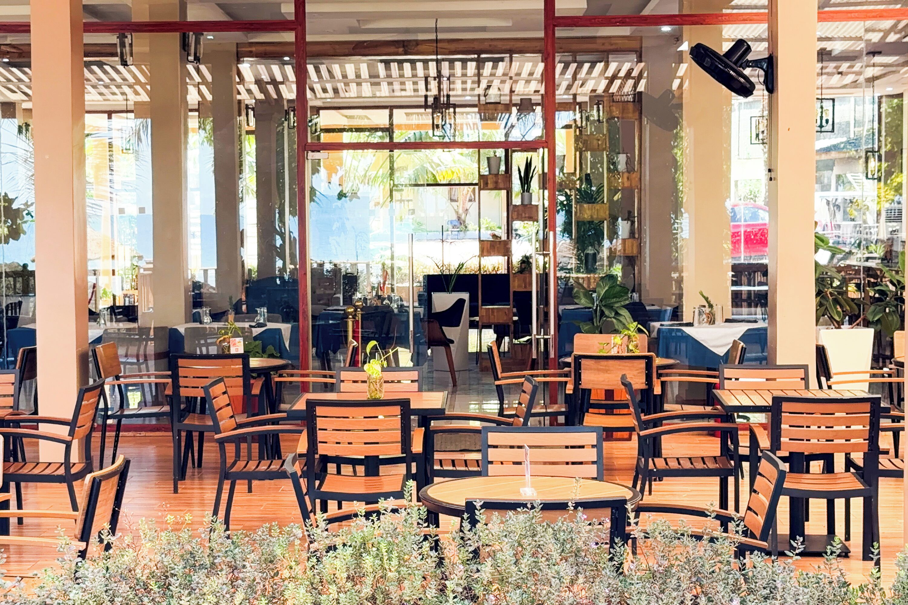 Un café al aire libre con mesas y sillas de madera, una zona de estar cubierta y macetas con plantas.