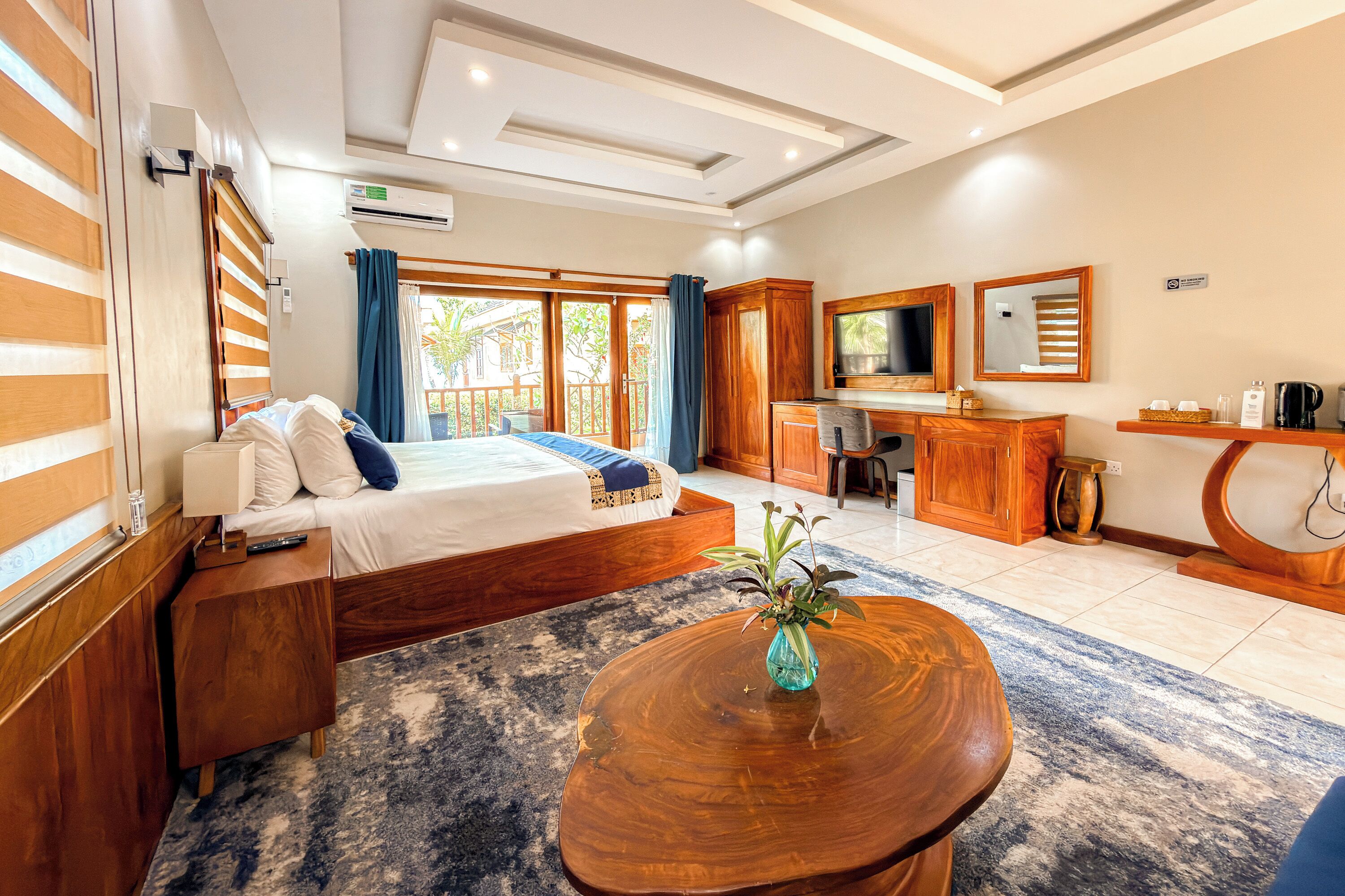 Habitación de hotel con cama, escritorio y zona de estar, que cuenta con un ventanal grande, aire acondicionado y una pequeña planta en la mesa.