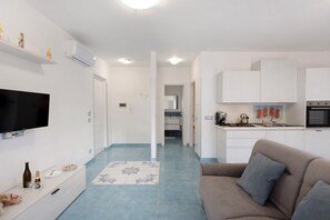 A modern living room with a sofa, a flat-screen TV, a kitchen area with cabinets and a stove, and a hallway leading to a bathroom.