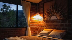 Una camera da letto con parete in mattoni, un'ampia finestra con tende e un letto con biancheria decorata, illuminata da una lampada fissata al muro.